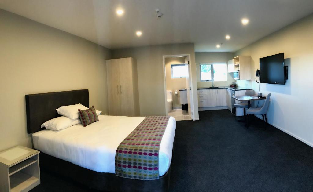 A spacious bedroom features a large bed adorned with a colorful throw blanket. A kitchenette is visible in the background, accompanied by a dining area that includes a small table and chair. Bright lighting from multiple sources enhances the welcoming ambiance.