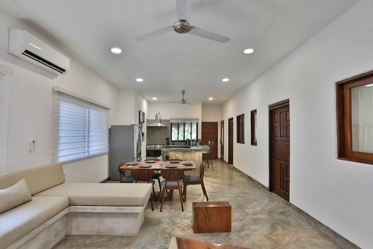 An open and airy living space is presented with a light-colored sofa and a wooden dining table set for six. A modern kitchen is equipped with stainless steel appliances, while multiple windows provide natural light, illuminating the neutral tones throughout the room.