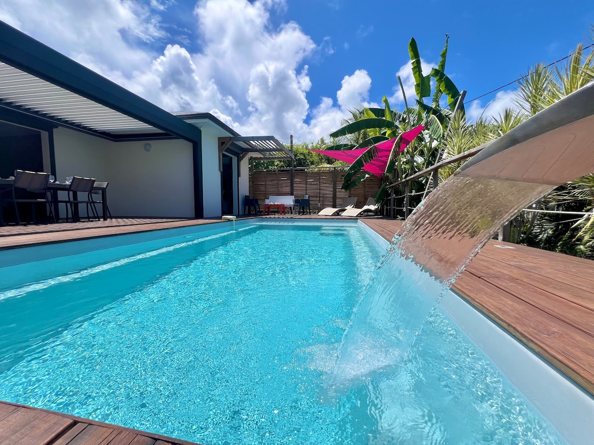 The swimming pool features a clear, inviting turquoise surface, complemented by a lively waterfall cascading gracefully from the edge. Surrounding the pool, a wooden deck is enhanced by tropical greenery, creating a serene outdoor space for relaxation and leisure.