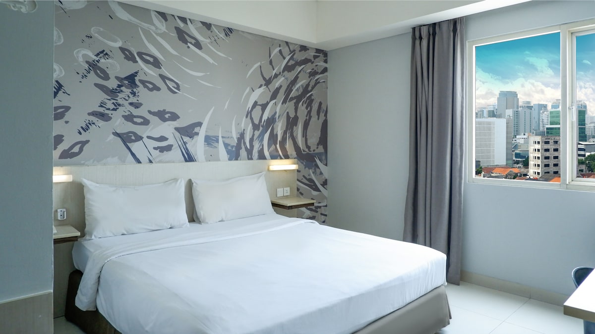A spacious sleeping area is adorned with abstract wall art, featuring a large bed dressed in crisp white linens. Natural light filters through a window, offering city views. Soft gray curtains frame the window, complementing the modern aesthetic.