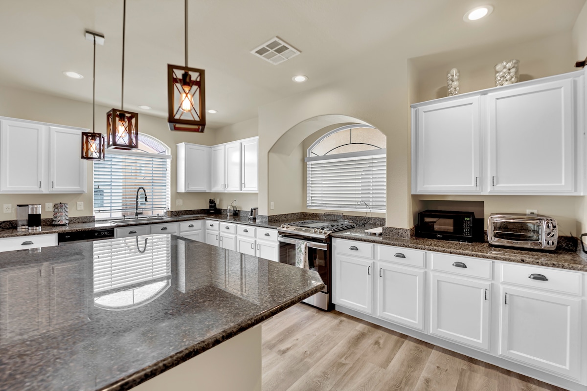 The kitchen showcases white cabinetry contrasted by dark granite countertops, with modern pendant lighting overhead. Stainless steel appliances, including a stove and microwave, are visible. Natural light filters through arched windows, and a spacious island provides additional counter space for meal preparation.