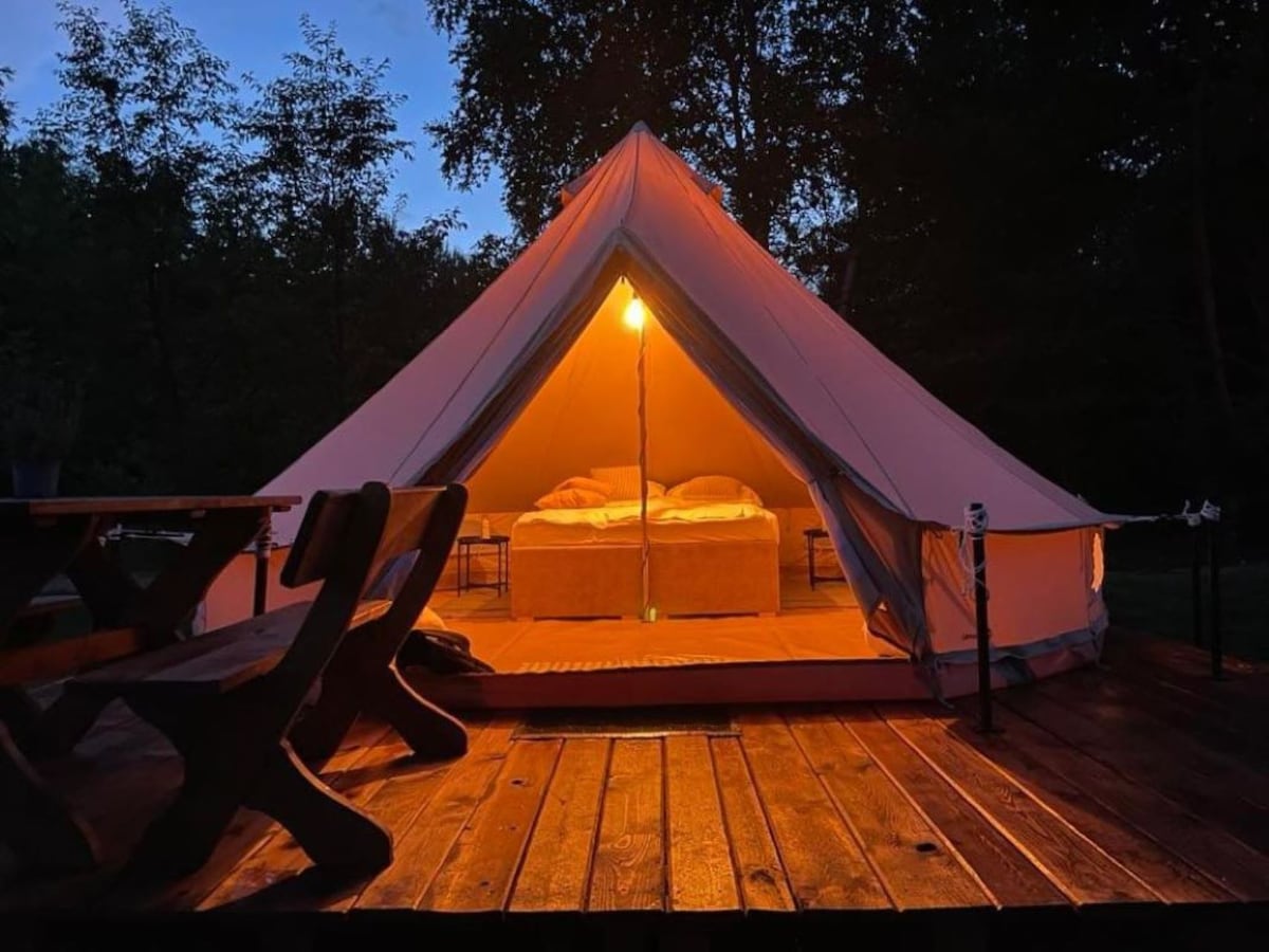A spacious glamping tent is illuminated by soft light, showcasing a large bed centered within. A wooden deck surrounds the tent, featuring a rustic dining set with several chairs. Trees frame the scene, enhancing the serene outdoor setting.