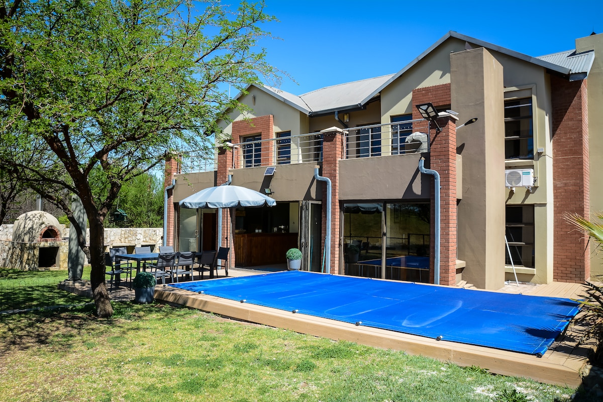 A modern house with large windows is set against a clear blue sky. An inviting outdoor space features a blue pool cover, a dining table with chairs, and an umbrella for shade. A stone pizza oven is visible in the background among greenery.