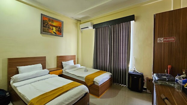 The room features two spacious beds with light-colored linens and decorative yellow throws. A large window is dressed with dark curtains, allowing natural light to filter in. A wooden wardrobe and a desk are positioned to provide convenient storage and workspace.