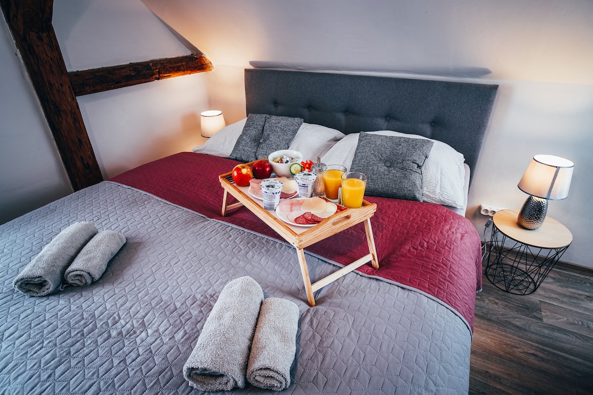 A spacious bed is dressed in a quilted grey and maroon cover, with a wooden breakfast tray featuring fruit, pastries, and beverages. Soft towels are neatly rolled at the foot of the bed, and bedside lamps provide gentle illumination.
