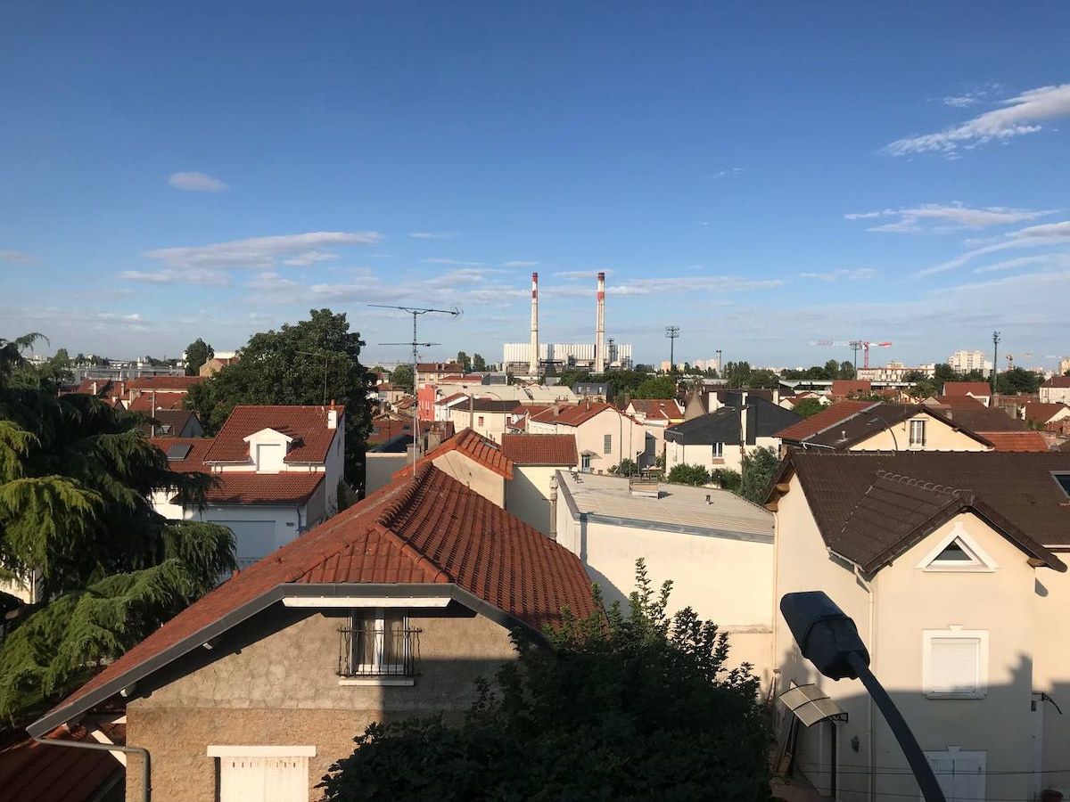 A view of a residential area is presented, showcasing a mixture of rooftops with varying colors and styles. In the distance, two tall structures are visible against a clear blue sky, while green trees and shrubs complement the scene.