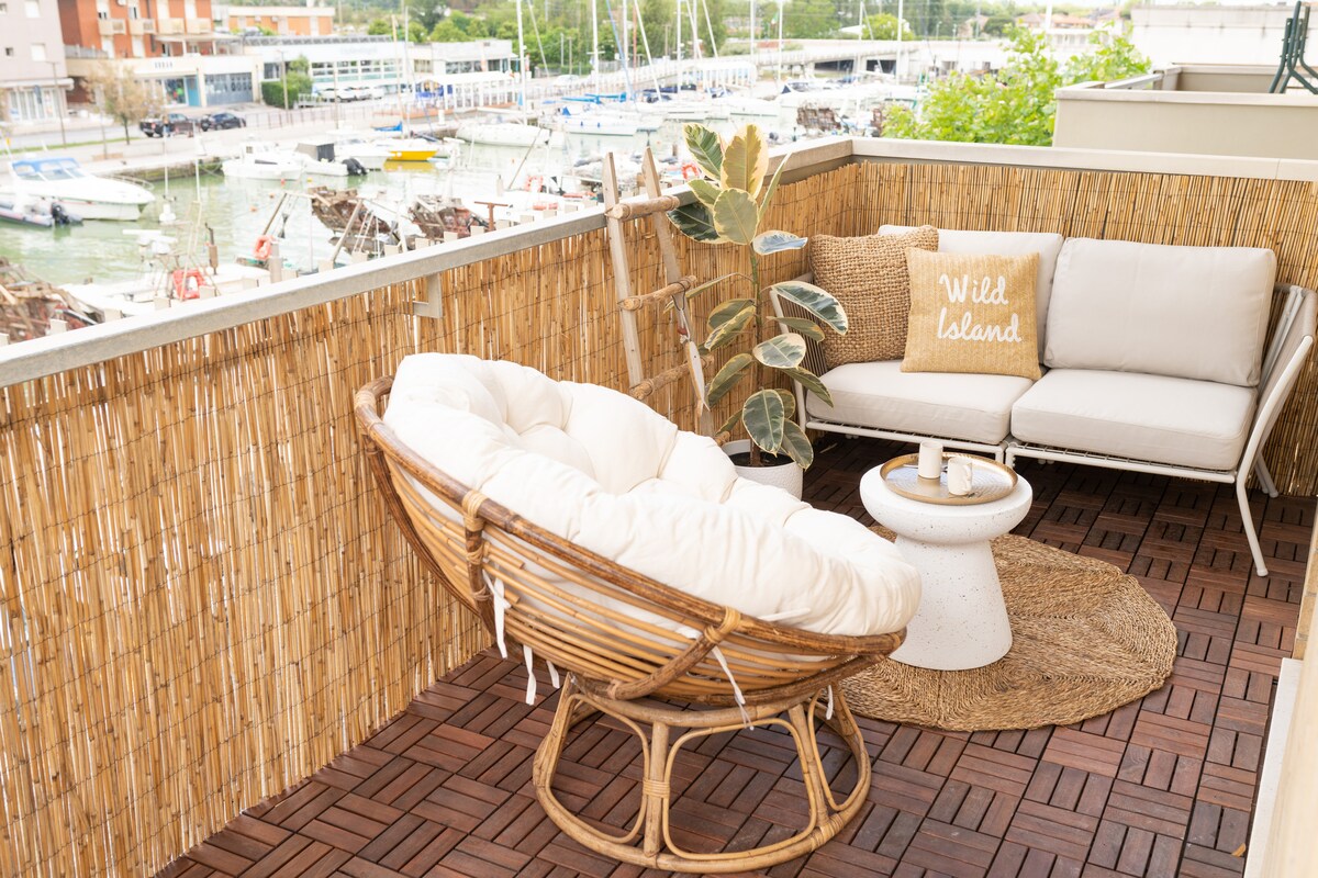 A spacious terrace features a comfortable seating area with a round wicker chair and a modern sofa, surrounded by a bamboo partition. A soft rug and a small table complement the setting, with views of boats and the harbor visible in the background.