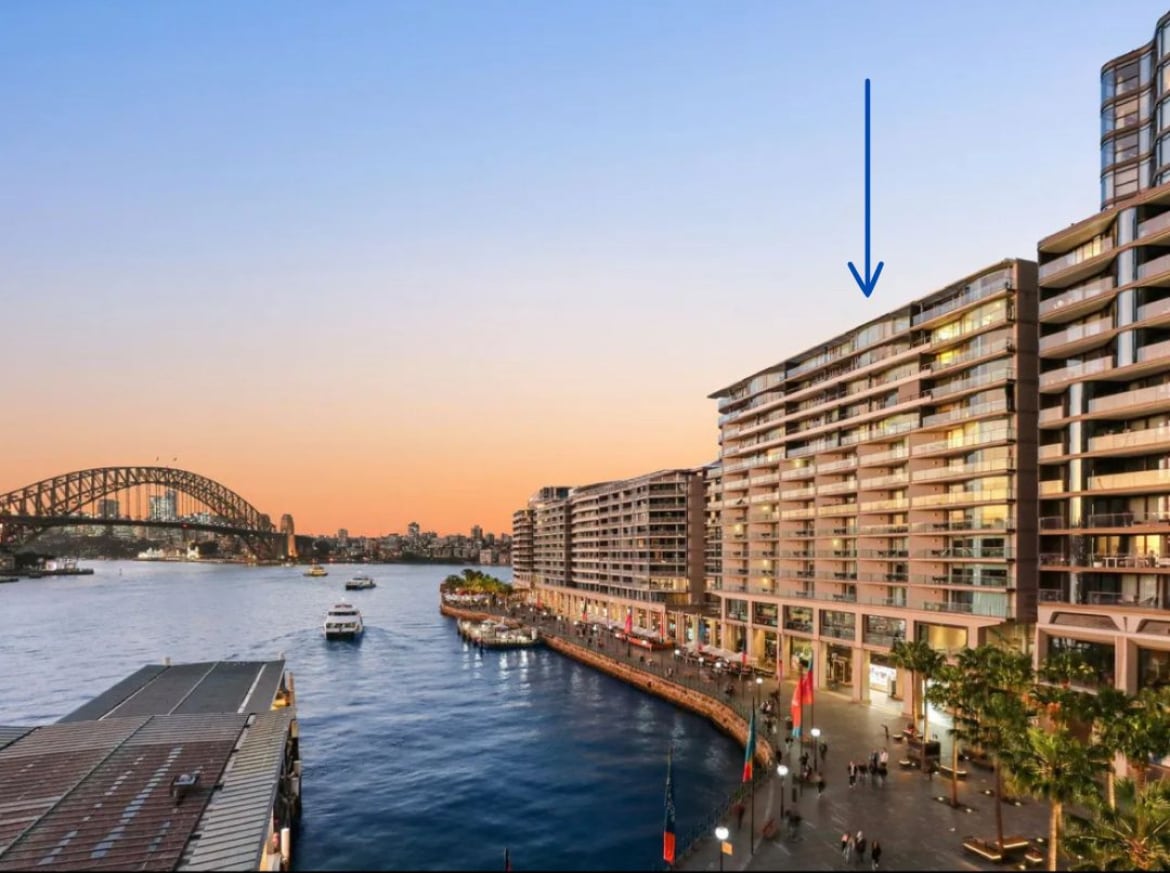 The image captures a scenic view of a waterfront area with a modern multi-story building highlighted by warm evening light. The Sydney Harbour Bridge is visible in the background, and boats can be seen navigating the water, emphasizing the vibrant atmosphere of the locale.