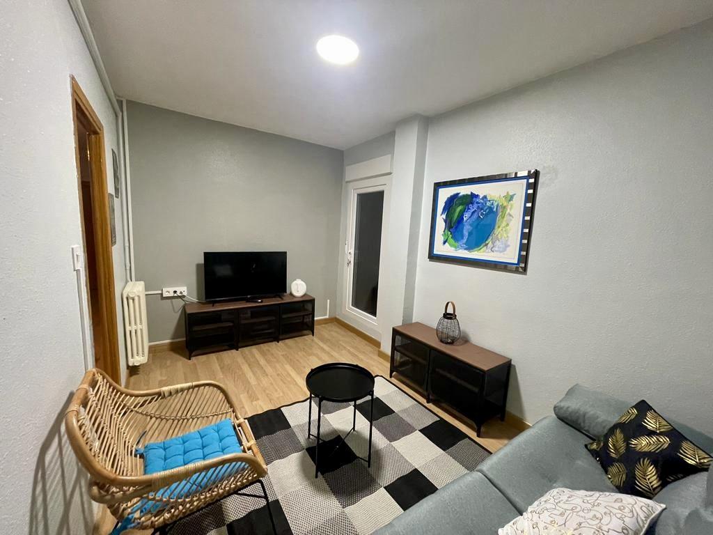 A comfortable living area features a light blue sofa and a woven chair, complemented by a small black coffee table. A patterned rug adds texture to the wooden floor. A television is positioned on a dark media unit, and a colorful wall art piece enhances the space.