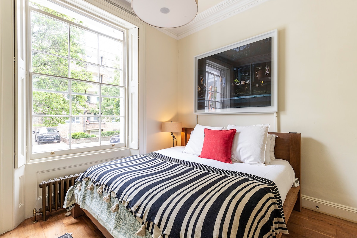 A bedroom features a spacious bed dressed in white linens and a striped blanket. A large window provides natural light, framed by elegant white trim. A piece of artwork hangs above the bed, while a bedside lamp adds warmth to the room.
