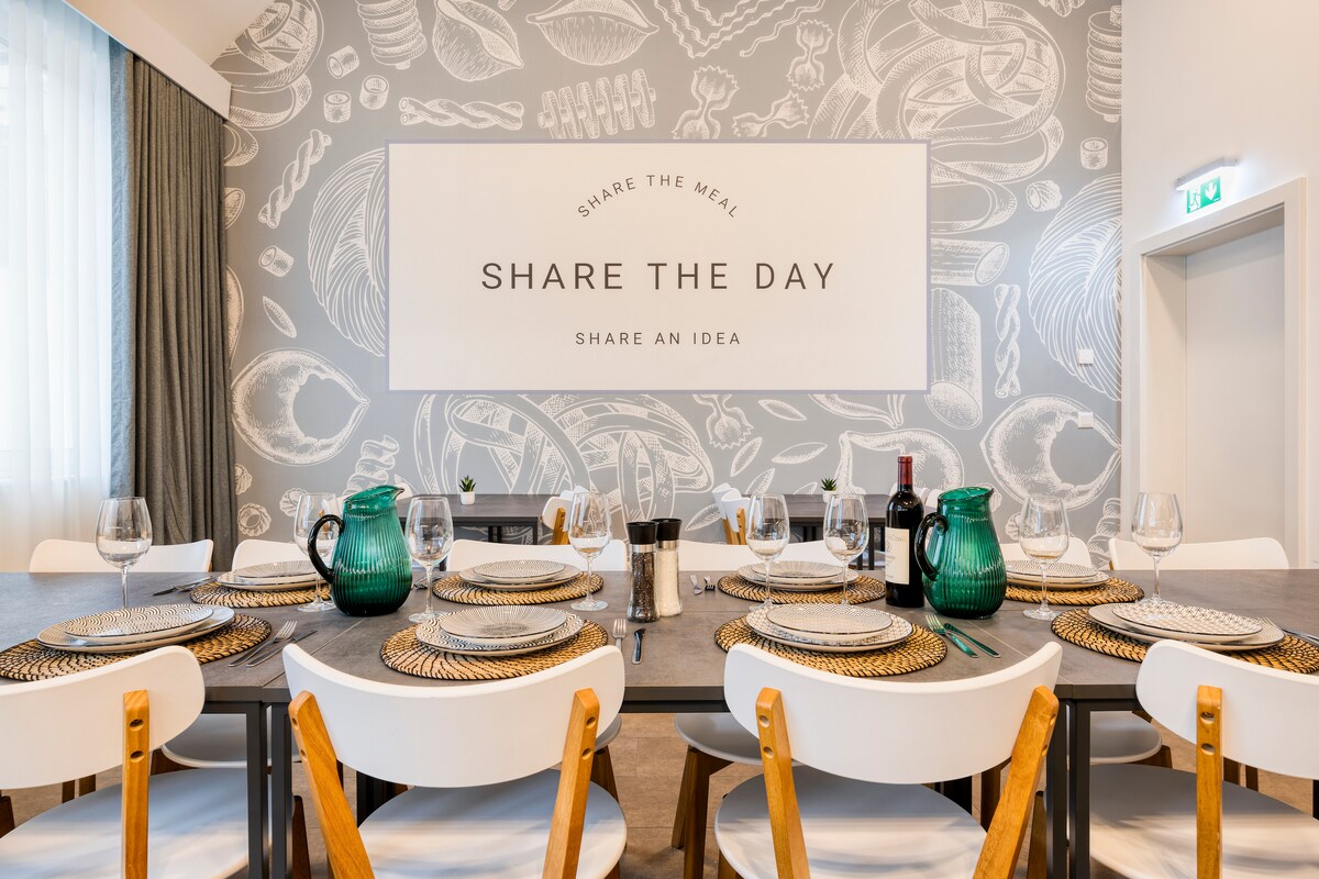 A communal dining area features a long table set for a meal, surrounded by white chairs with wooden legs. The wall showcases a decorative mural of food items, complemented by a large sign encouraging sharing. Elegant table settings include green glassware and woven placemats.