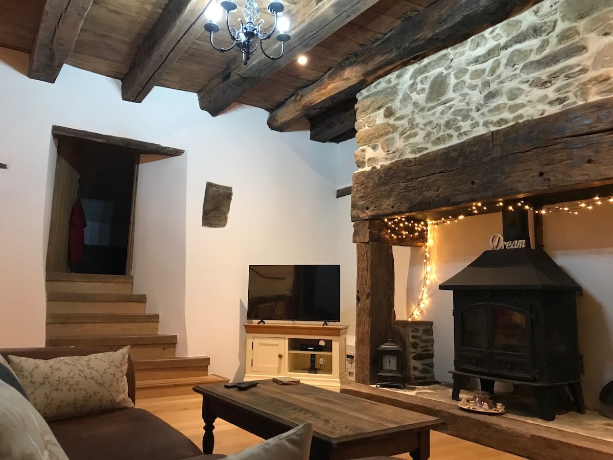 The lounge area features exposed wooden beams and a stone fireplace, complemented by a rustic coffee table. A television is positioned in front of a cozy seating arrangement. Soft lighting is provided by a chandelier and decorative string lights along the fireplace.