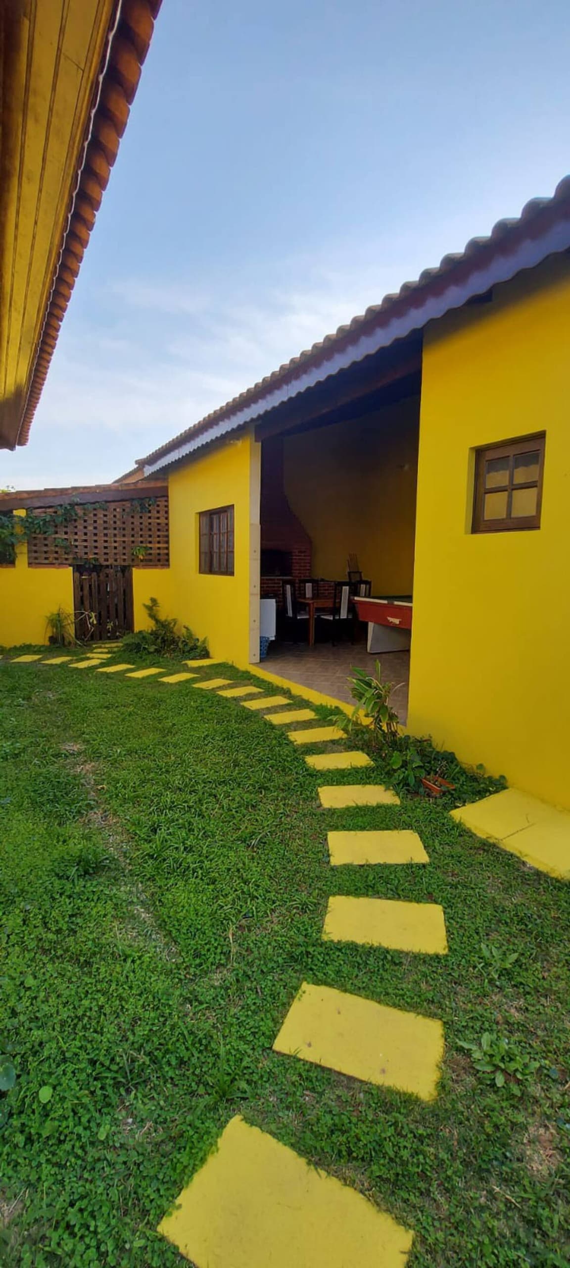 The exterior of the house features a vibrant yellow façade, with a well-maintained garden and a stone pathway leading to an open area. The entrance to the kitchen is visible, showcasing a glimpse of indoor seating and appliances.