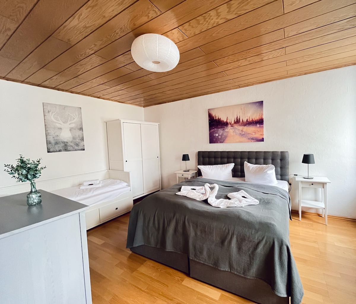 A spacious bedroom features a large boxspring bed with crisp linens and two bedside tables, each holding a lamp. A light-colored wardrobe is positioned beside a smaller pull-out bed, while art pieces with nature scenes adorn the walls. Natural light enters through a window.
