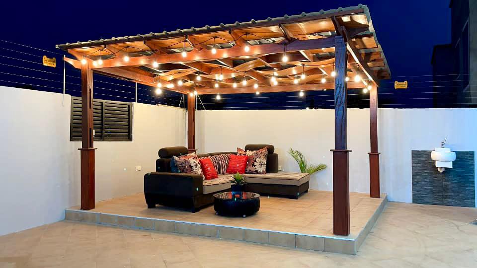 A shaded pergola offers a cozy seating area adorned with cushions in rich colors. Soft glow lights hang above, enhancing the inviting atmosphere. The patio is paved with smooth tiles, surrounded by simple greenery and a wall feature, creating a peaceful outdoor retreat.
