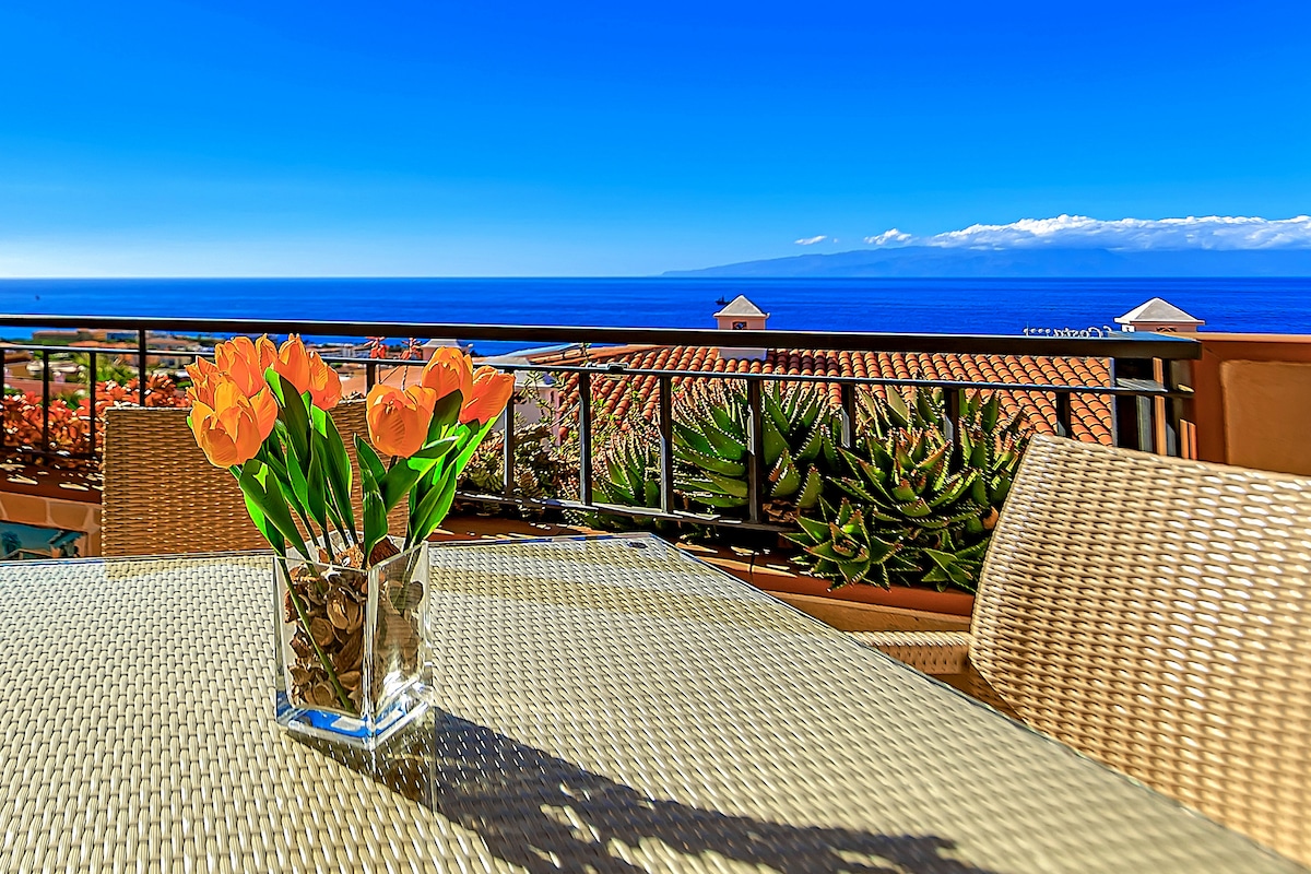 A glass vase filled with orange tulips is placed on a woven table. Lush greenery and colorful plants are visible in the background, with a clear view of the ocean and distant mountains under a blue sky.