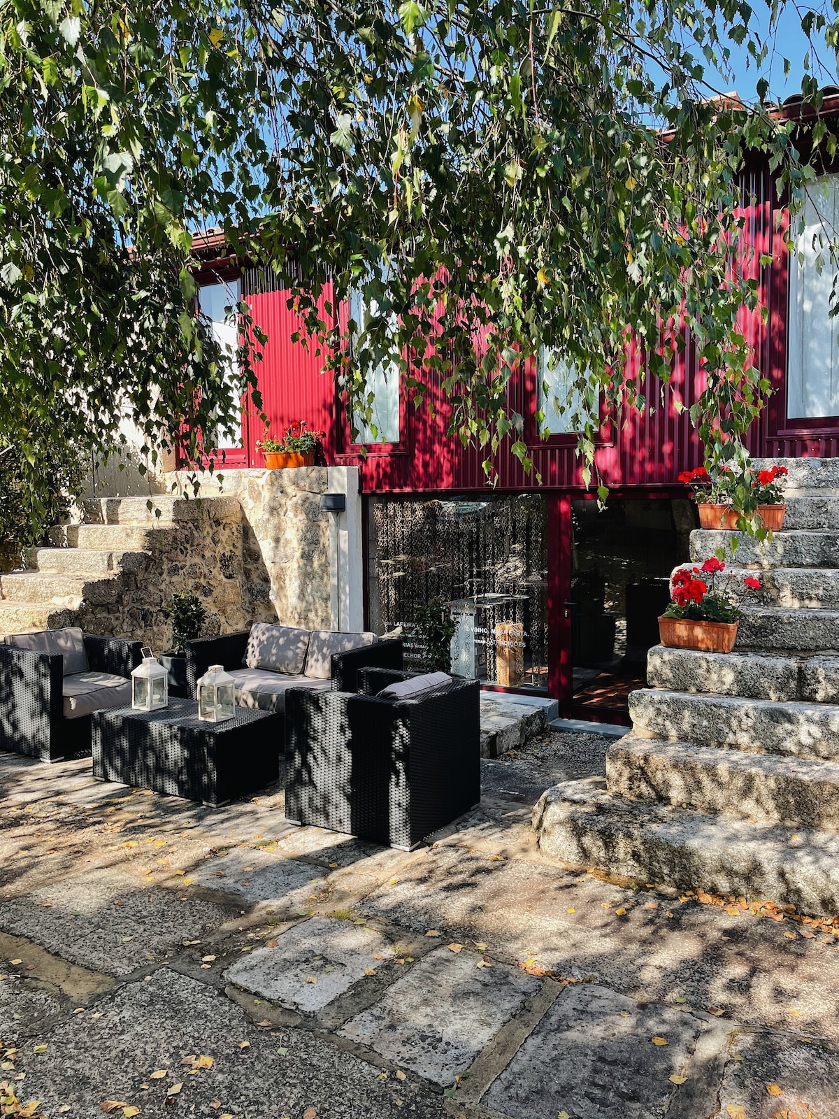 An outdoor seating area is presented, featuring a set of dark furniture arranged around a low table. The space is framed by stone steps leading up to a red structure, with potted flowers enhancing the inviting atmosphere. Natural light filters through the leafy trees above.