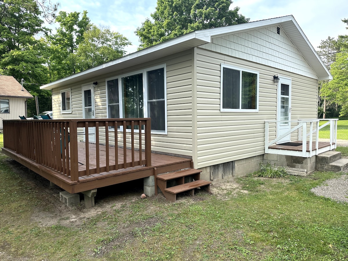 A modern cabin exterior features a spacious wooden deck with steps leading to the entrance. Large windows allow for natural light, and the surrounding greenery creates a serene setting. The cabin is situated on a grassy area, emphasizing its connection to nature.