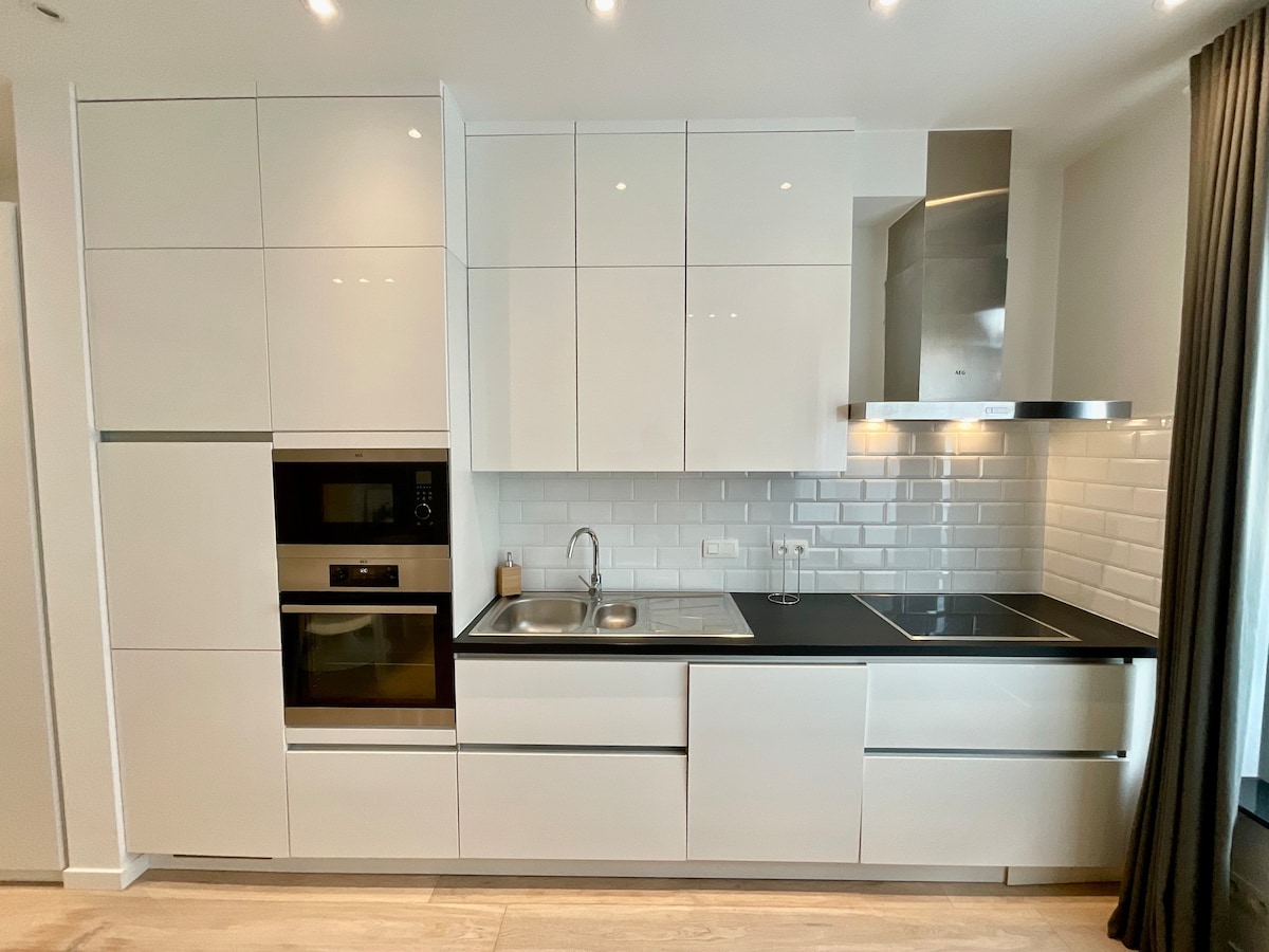 A modern kitchen layout is presented with glossy white cabinetry and a sleek black countertop. Stainless steel appliances, including an oven and induction cooktop, are integrated into the design. A stainless steel sink is positioned under upper cabinets, with minimalistic tile backsplash providing a clean aesthetic.