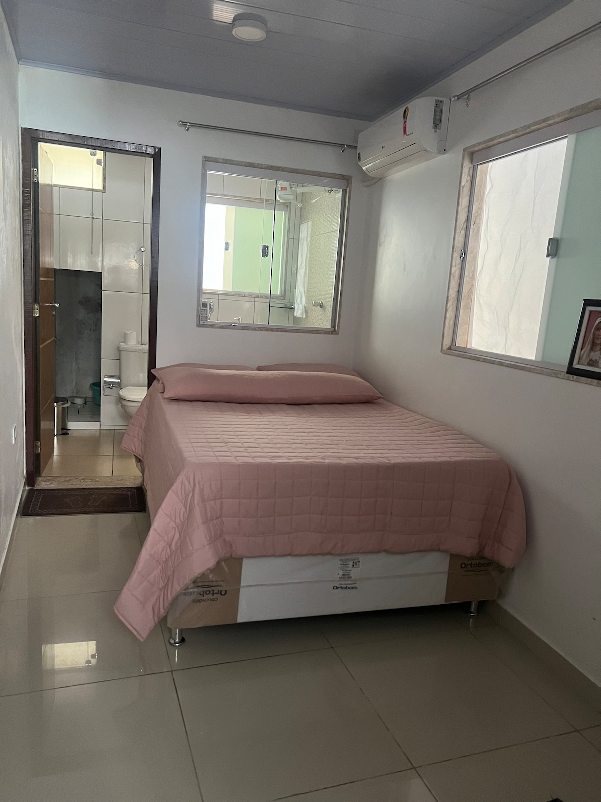 A cozy bedroom is presented, featuring a spacious bed dressed in a soft pink coverlet. Natural light enters through two windows, illuminating the room's simple decor. An adjoining bathroom is visible in the background, with tiled walls and modern fixtures.
