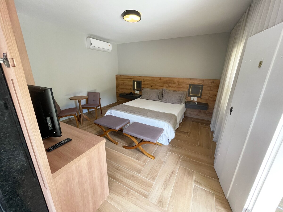 The room features a spacious bed against a wooden wall with two modern bedside tables. A small dining area with a table and two chairs occupies one corner. A sleek TV is mounted on a wooden cabinet, and natural light filters through sheer curtains.