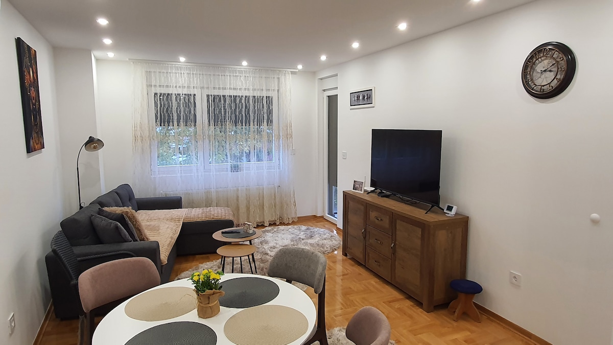 A cozy living area is shown, featuring a comfortable sectional sofa, a modern coffee table, and a flat-screen television mounted on a wooden console. Natural light filters through sheer curtains, enhancing the warm wood flooring and neutral-colored walls.