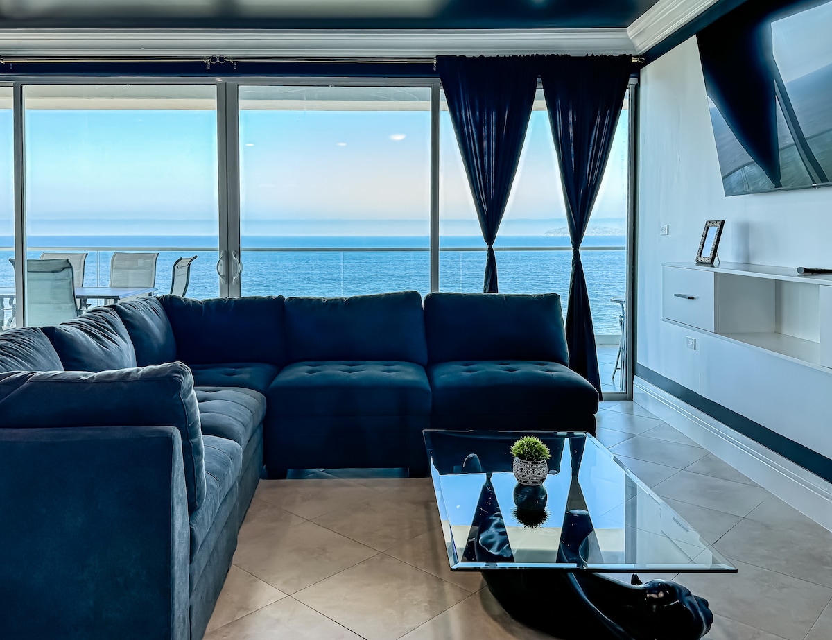 A spacious living area features a large sectional sofa in a deep blue hue, complemented by a glass coffee table. Expansive floor-to-ceiling windows offer unobstructed views of the Pacific Ocean, with soft natural light brightening the room. Dark curtains frame the windows, enhancing the serene atmosphere.