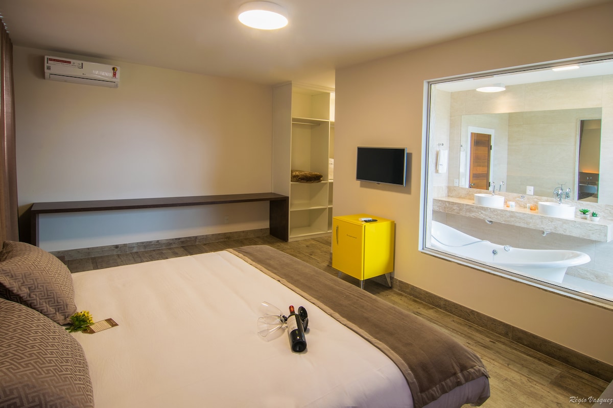 A spacious bedroom is presented, featuring a comfortable king-sized bed with neutral bedding. A large window frames a view of a modern bathroom with a freestanding bathtub. A flat-screen TV is mounted on the wall, and a contemporary yellow cabinet adds a pop of color.