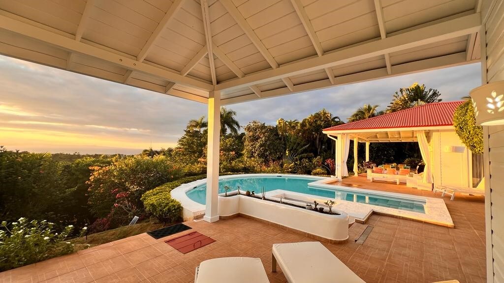 A serene outdoor area features a swimming pool surrounded by lush greenery. Lounge chairs are positioned near the pool, while a covered seating area showcases comfortable furniture. The vibrant landscape is complemented by a colorful sunset in the background.