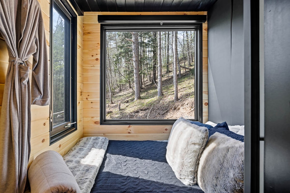 A cozy sleeping area is visible, featuring large windows that frame views of the surrounding woods. Soft blankets and decorative pillows are arranged on a spacious bed, with natural wood walls creating a warm atmosphere. Light streams in, enhancing the serene environment.