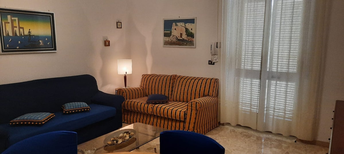 A spacious living room is presented with two comfortable sofas, one in a solid blue hue and the other striped in warm tones. A glass coffee table occupies the center, complemented by decorative items. Natural light filters through sheer curtains, illuminating the room.