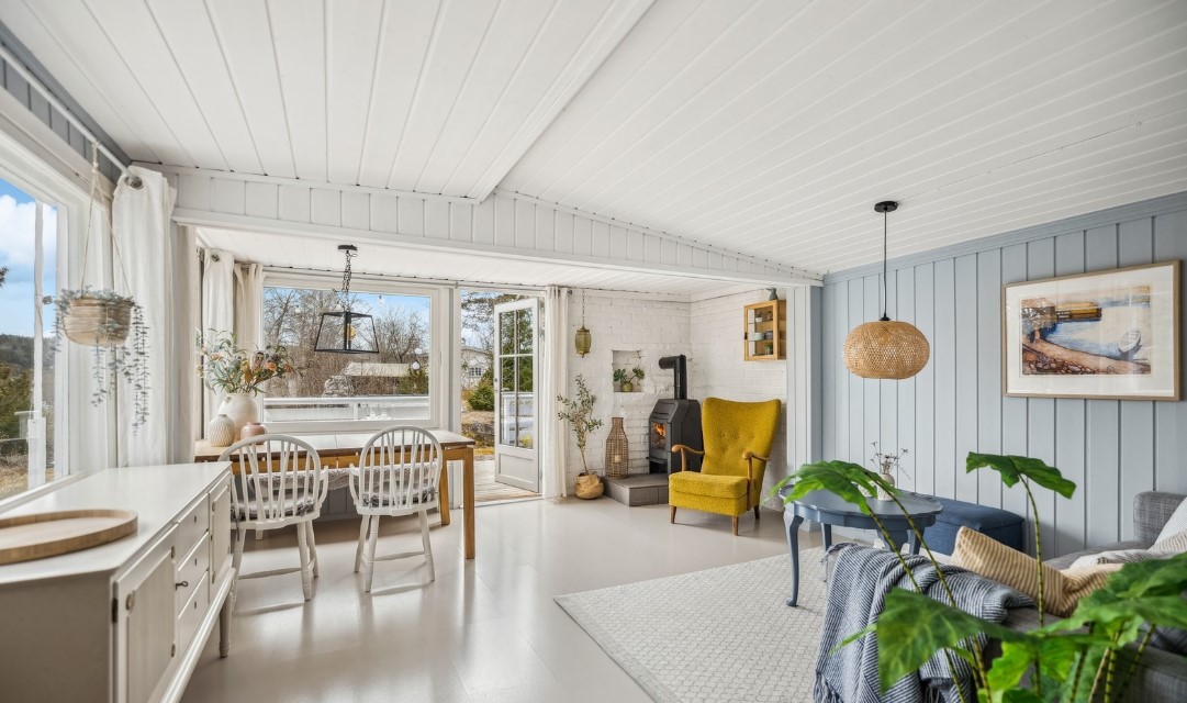 A bright living area features large windows allowing natural light to fill the space. A wooden dining table set for six is positioned alongside a comfortable yellow armchair and a cozy gray sofa. Elements of nature are included with potted plants throughout.