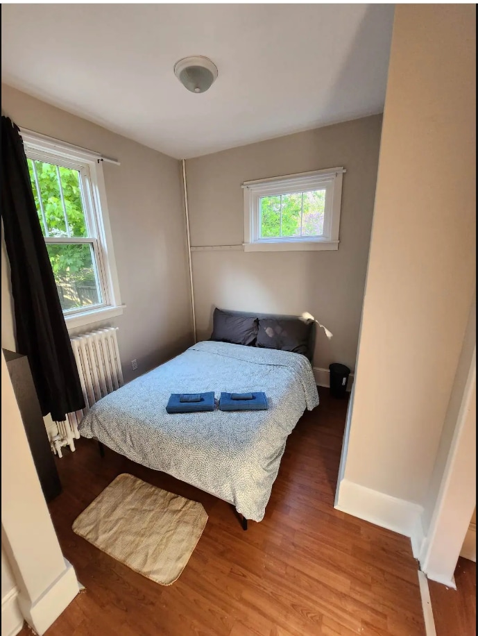 A cozy bedroom features a spacious bed with a patterned blanket, complemented by two neatly arranged towels on top. Natural light enters through the small window, illuminating the warm wood flooring. A soft area rug lies at the foot of the bed, enhancing the inviting space.