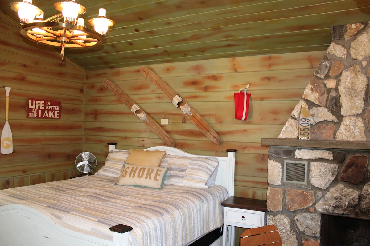 A cozy bedroom features a spacious bed with a striped blanket, accompanied by a decorative pillow that reads 'SHORE.' Rustic wooden walls are paired with stone accents, including a fireplace. Nautical decor adds character, while a fan provides comfort.