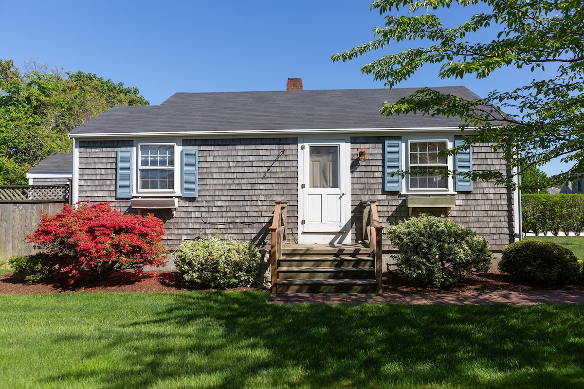 A charming cottage features a grey shingled facade with blue shutters. A wooden staircase leads to the front door, surrounded by lush greenery and colorful flower beds. A well-manicured lawn extends in front, enhancing the welcoming appearance of the home.