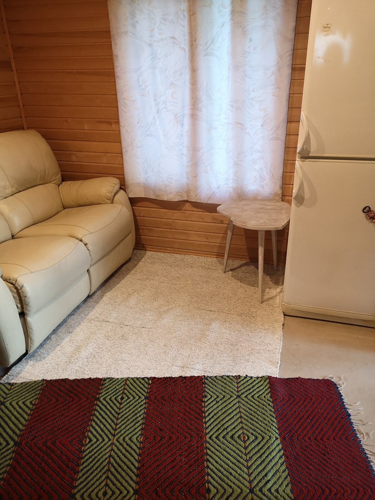 A small seating area is highlighted by a light-colored sofa and a circular side table. A window covered with light fabric allows natural light to filter in, illuminating the space. A patterned area rug adds color to the floor, complementing the wooden walls.