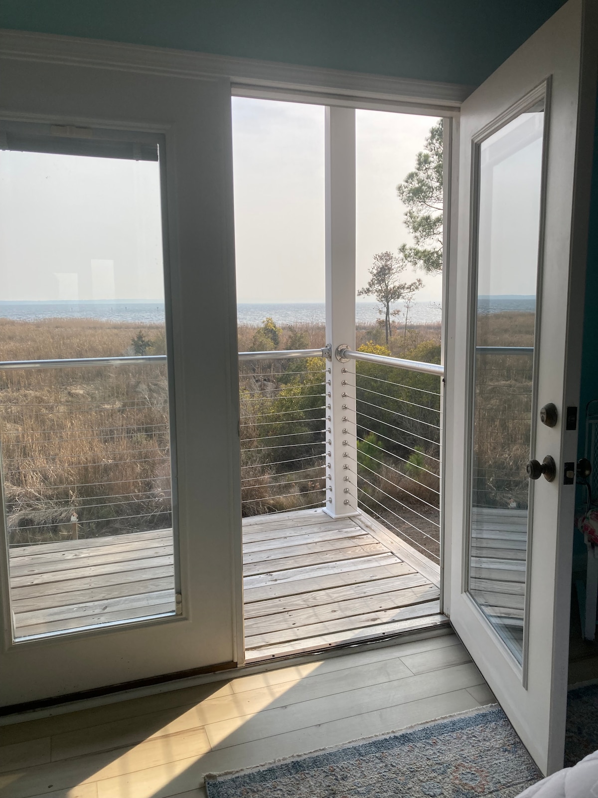 A view of the outdoor balcony is framed by glass doors, offering a glimpse of the expansive landscape that includes both trees and water. Natural light is evident in the space, enhancing the connection to the outdoors.