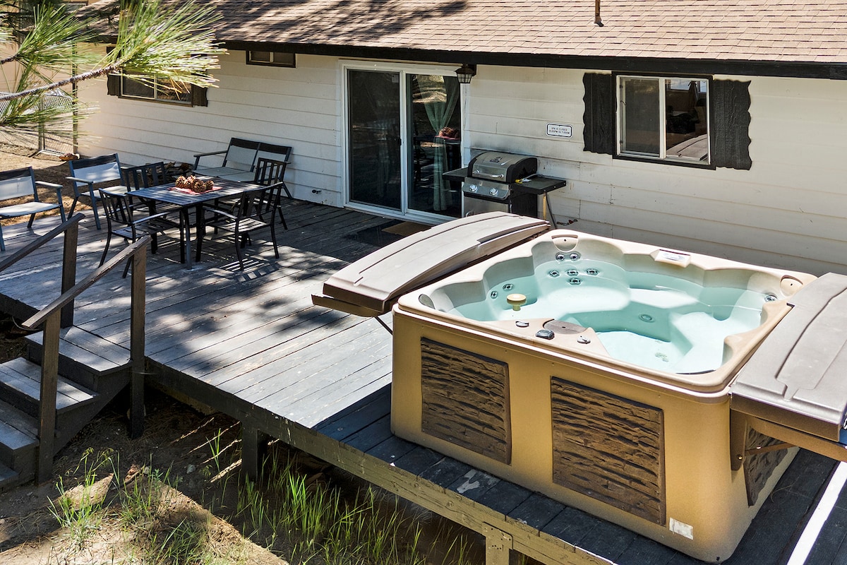 A spacious deck features a hot tub and a dining area with a table and chairs, providing a setting for outdoor meals and relaxation. A barbecue grill is also visible, making it convenient for outdoor cooking. Lush greenery surrounds the area, enhancing the natural ambiance.