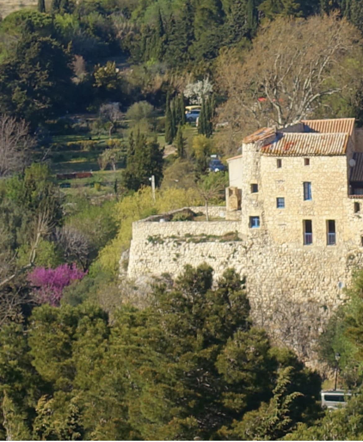 The image showcases a stone house perched on a rocky outcrop, surrounded by lush greenery. The structure reflects rustic architecture with a terracotta roof. Colorful trees and greenery create a vibrant backdrop, emphasizing the natural beauty of the setting.