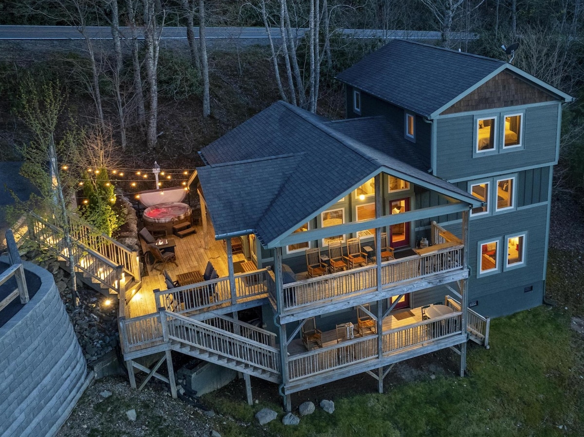 A three-story chalet is highlighted by a spacious deck featuring seating and a hot tub, surrounded by warm outdoor lighting. The structure's design showcases large windows, and a well-manicured lawn is visible, enhancing the connection to nature and creating an inviting outdoor space.