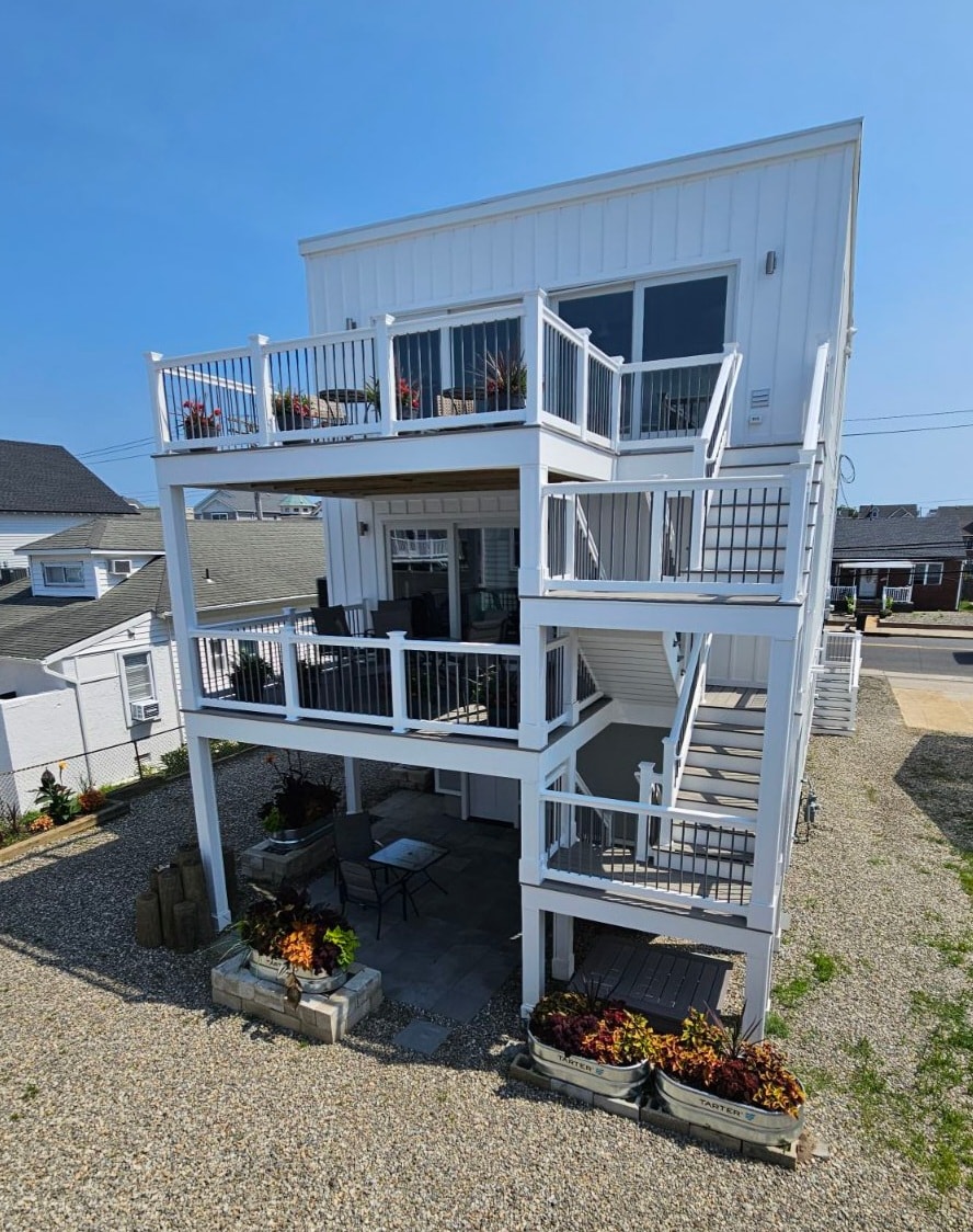 The newly built house features three levels, each with a spacious deck surrounded by white railings. Potted plants adorn the decks, adding greenery. The ground level has a paved area with a small table and chairs, creating a welcoming outdoor space. Natural light illuminates the exterior.