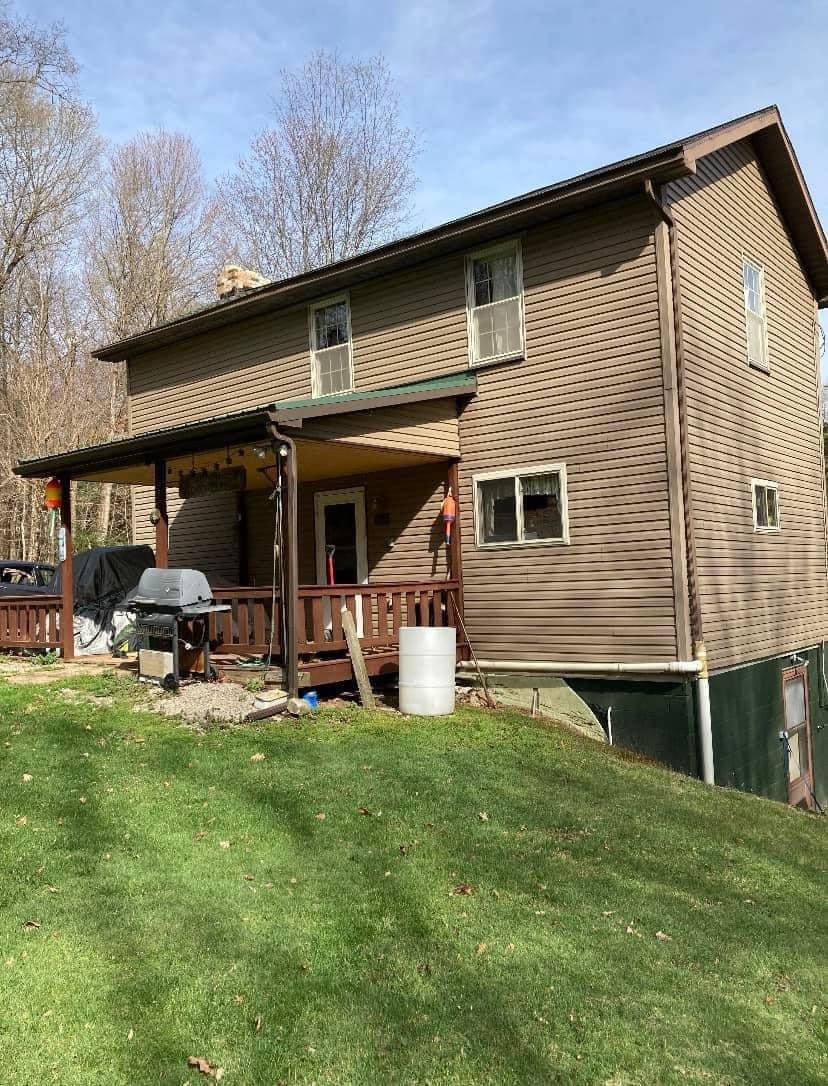 A two-story exterior of the house is shown, featuring wooden siding and a covered porch. The porch is furnished with a barbecue grill. The surrounding landscape includes a grassy lawn and trees in the background, indicating a natural setting.