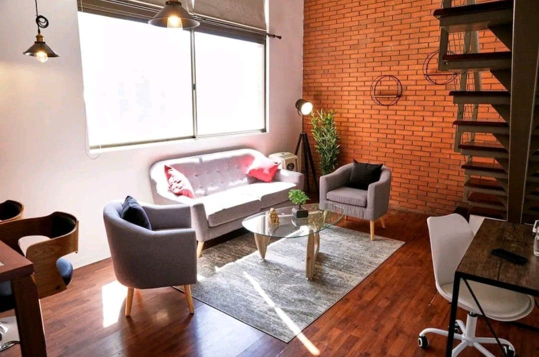 A modern living area features a light grey sofa and two grey armchairs arranged around a glass coffee table. A textured rug anchors the seating area on warm wood flooring. Natural light streams in through a large window, illuminating the exposed brick wall and indoor plant.