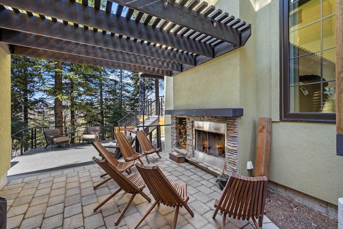 An outdoor covered patio features multiple wooden lounge chairs arranged around a stone fireplace. The space is shaded by a wooden pergola and is surrounded by trees, creating a serene environment suitable for relaxation.