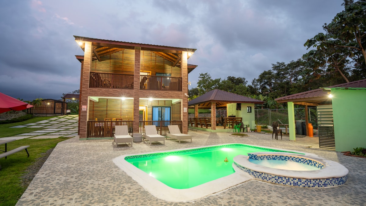 A multi-story cabin surrounded by greenery features a well-lit exterior. The swimming pool is enhanced by a small jacuzzi, while lounge chairs are positioned beside the water. A shaded area with outdoor seating is visible, promoting relaxation in a natural setting.