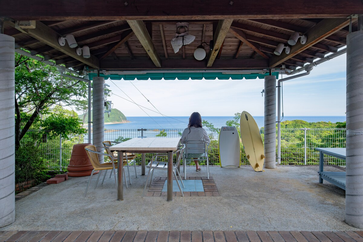 An outdoor covered pavilion overlooks a scenic ocean view, featuring a wooden dining table surrounded by several chairs. A surfboard leans against the wall, and a quiet individual is seated, enjoying the tranquil atmosphere of nature.