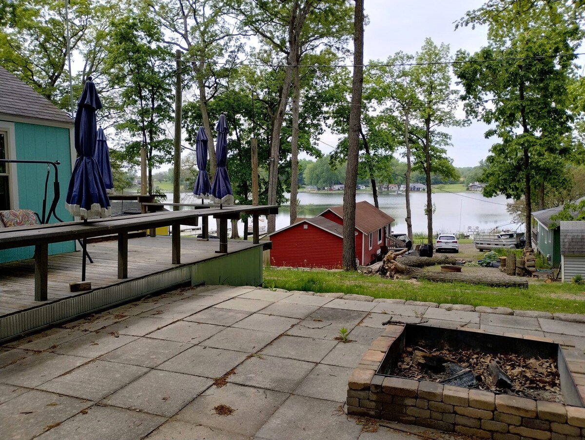 A spacious outdoor patio features a stone fire pit surrounded by laid stone tiles. A long wooden table is set with several umbrellas for shade, overlooking a tranquil lake lined with trees. Colorful cabins are visible in the background, contributing to the serene environment.
