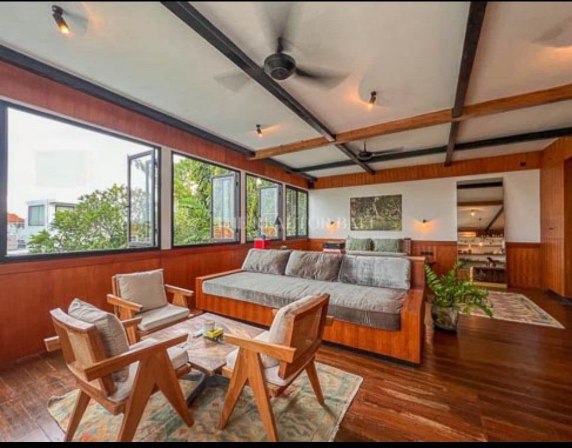 A spacious lounge area is designed with wood accents and large windows that invite natural light. Comfortable seating is provided by a large daybed and two wooden armchairs, complemented by a coffee table and decorative rug. The open layout offers views into the kitchen area.