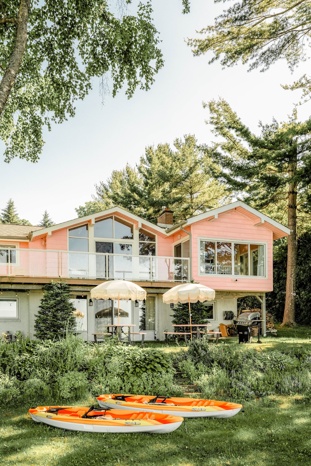 The exterior of the cottage features a charming pink facade with large windows allowing for ample natural light. Two kayaks rest on the grassy area below, accompanied by beach umbrellas providing shade. Tall trees surround the property, enhancing the serene lakeside environment.