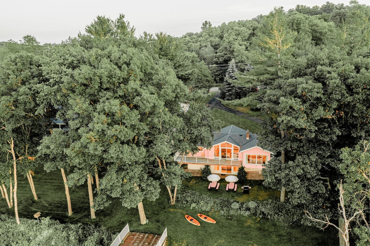 An aerial view captures a charming cottage nestled among tall trees, with a spacious deck. Two beach umbrellas and pink picnic tables are visible in the yard, alongside two kayaks resting on the grass, creating a serene outdoor setting.